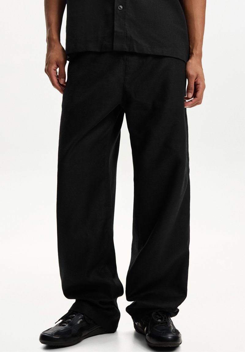 Personne portant un pantalon noir ample, une chemise noire boutonnée et des baskets noires, debout sur un fond blanc.