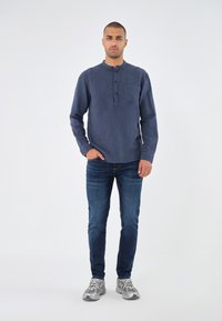 Marineblaues Langarmhemd mit Rundkragen, fünf Knöpfen in der Knopfleiste und Brusttasche, kombiniert mit blauen Jeans und grauen Sneakers.