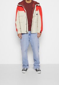 Vaaleanbeige ja punainen huppari, jossa kaksi etutaskua, yhdistetty viininpunaiseen paitaan ja sinisiin farkkuihin, viimeistelty mustilla lenkkareilla.