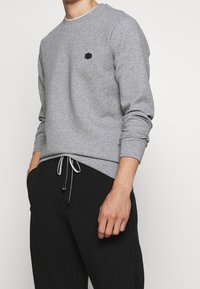 Grå sweatshirt med långa ärmar, rundad halsringning och en svart hexagonal logotyp på bröstet. Matchad med svarta byxor med snörning.