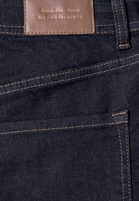 Poche de jean en denim bleu foncé avec des coutures marron et une étiquette en cuir marron cousue au-dessus portant l'inscription "Street One Denim All Time Favourite."