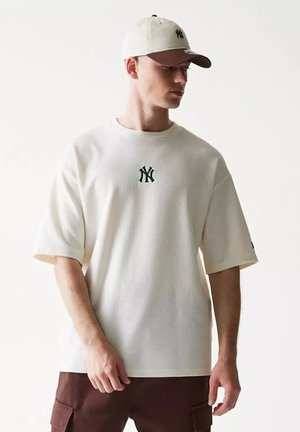 Giovane uomo che indossa un cappellino beige e una camicia beige oversize con un logo marrone scuro, abbinati a pantaloni cargo marrone scuro su uno sfondo semplice.