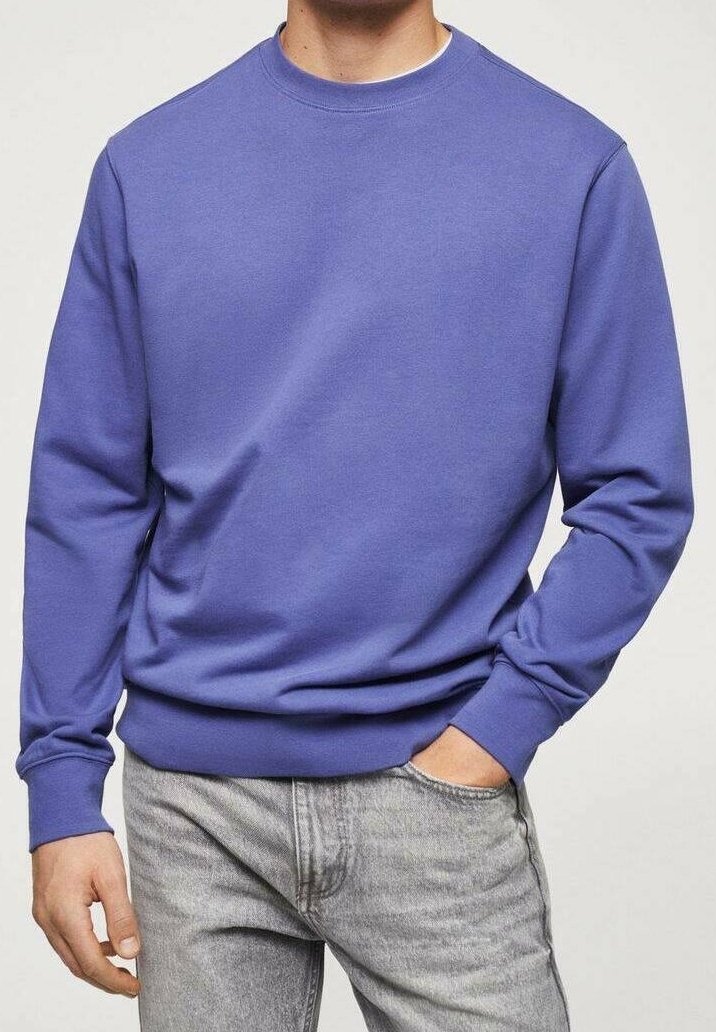 Homme portant un sweat-shirt violet uni à col rond et un jean gris clair, une main dans la poche et l'autre détendue sur le côté.