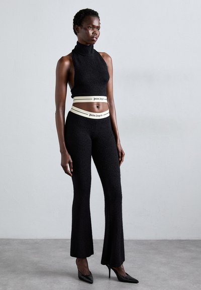 Woman wearing black sleeveless crop top and matching flared pants with white waistband logos, standing on gray floor against plain wall.