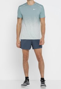 Nike sportstrøje i gradient teal med korte ærmer og reflekterende detaljer, kombineret med marineblå shorts. Det samlede design er sportsligt og funktionelt.