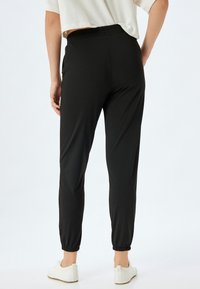 Joggers negros con cintura elástica y perneras estrechas, que presentan puños en los tobillos, una textura suave y un diseño minimalista, combinados con zapatos blancos.
