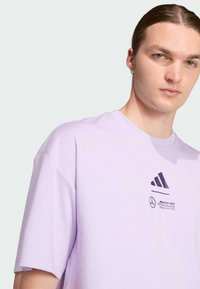 Camiseta de manga corta en color lavanda, hecha de algodón, con un logo impreso en púrpura oscuro. Diseño de cuello redondo y corte relajado.