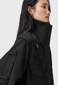 Cappotto nero con colletto alto, cerniera laterale e tasche applicate. Il materiale sembra strutturato con una finitura opaca. Gli accessori includono spalline.