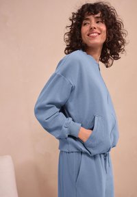 Svetlo modra zip-up pulover s mehko teksturo, zaobljenim ovratnikom in stranskimi žepi. Ima sproščen kroj in rebraste manšete.