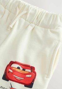 Pantalones de chándal de color crema hechos de una tela suave que presentan un gráfico de un coche de dibujos animados rojo en la parte frontal, con una cintura elástica y cordón.