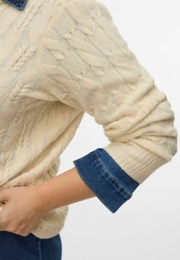 Suéter de punto trenzado crema con puños de mezclilla enrollados, con un patrón texturizado y mangas largas, parcialmente visible sobre un fondo neutro.