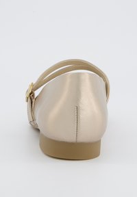 Zapato plano metálico dorado con una correa y detalle de hebilla, punta redondeada, textura suave y suela baja color marrón. Vista trasera mostrada.