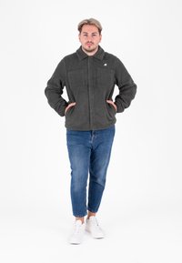 Giacca in pile verde scuro con colletto, tasche anteriori e maniche lunghe, abbinata a jeans blu e sneakers bianche.