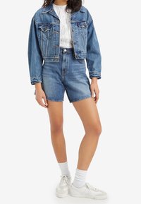 Džínová bunda má klasický střih, světle modrou barvu a přední kapsy. Kombinována s džínovými kraťasy s vysokým pasem a bílými teniskami.