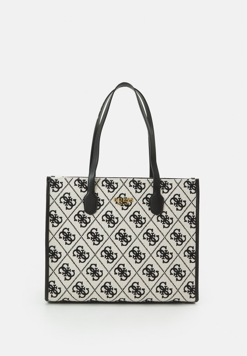 Rechthoekige beige draagtas met zwarte handgrepen, voorzien van een herhalend zwart monogrampatroon en een klein gouden "GUESS EST. 1981"-logo aan de voorkant.