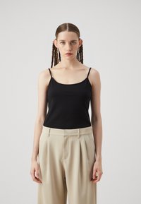 Black sleeveless top with thin straps, made of smooth fabric; paired with light beige high-waisted trousers featuring pleats.