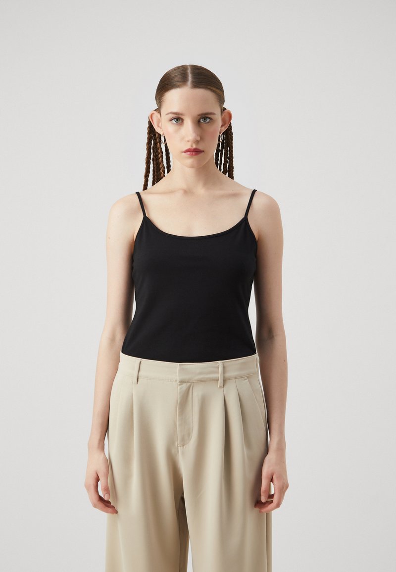 Black sleeveless top with thin straps, made of smooth fabric; paired with light beige high-waisted trousers featuring pleats.