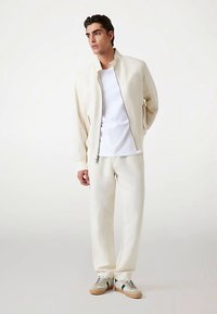 Lysecremefarvet let jakke med lynlås og sidelommer, kombineret med matchende sweatpants og sneakers i neutrale toner. Enkel design.