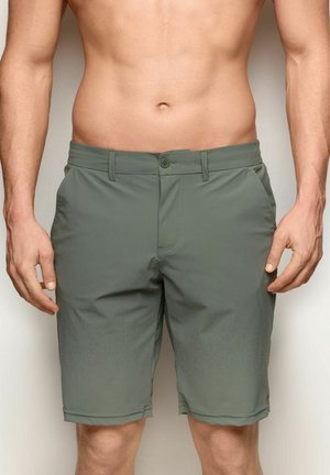 Oberkörperfreier Mann trägt olivgrüne knielange Shorts mit Frontknöpfen und Seitentaschen, steht vor einem neutralen Hintergrund.