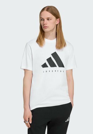 Joven con cabello largo que lleva una camiseta blanca con el logo de la Juventus y pantalones negros, de pie con una mano en el bolsillo.