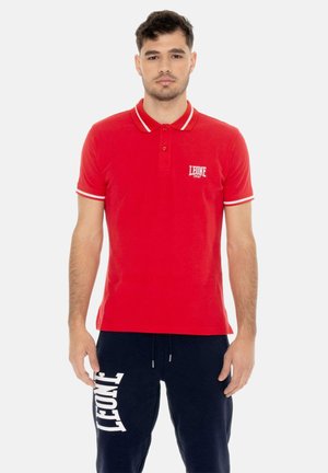 Homme portant un polo rouge avec des bordures blanches et un logo, associé à un pantalon bleu marine avec un grand logo blanc sur la jambe gauche.