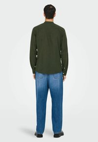 Chemise à manches longues de couleur vert foncé avec une coupe droite et des poignets boutonnés, associée à un jean bleu clair à jambes larges. Des chaussures noires complètent le look.