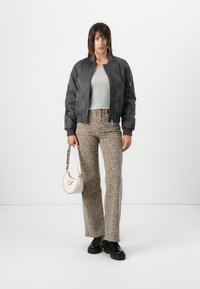 Chaqueta bomber negra con detalles bordados, top gris ajustado, pantalones acampanados de estampado de leopardo y zapatos de plataforma negros, sosteniendo un pequeño bolso blanco.