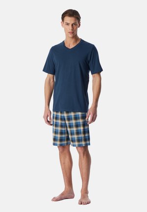 Man standing barefoot wearing a navy blue short sleeve shirt and blue, white, and brown plaid shorts on a plain background.