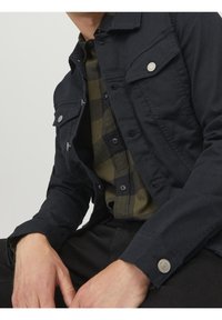 Mörk denimjacka med fyra framfickor, silverdetaljer och knäppta ärmslut, lager på en grön och svart rutig skjorta.