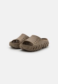 Brown slide sandals made of textured material, featuring an open toe, ventilation holes on the upper, and a wavy, contoured sole design.