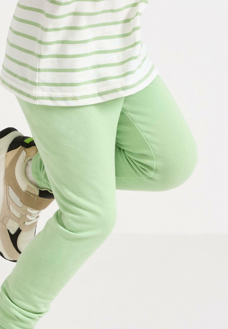 Lysegrønne leggings i blødt stof med en glat tekstur, båret med en hvid og grøn stribet skjorte og kraftige hvide sneakers.