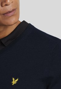 Navy knit sweater with a round neckline and black collar; features a small yellow eagle emblem on the chest. Smooth texture.