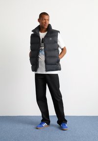 Sort puffer vest, med lynlås fortil, høj krave. Båret over en hvid T-shirt med sort og blå logo. Sorte bukser og blå Adidas-sko.