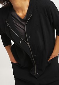 Svart långärmad topp med en hel dragkedja fram, tryckknappar och en avslappnad passform, tillverkad i ett lätt texturerat tyg.