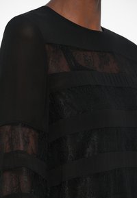 Svart kjole med transparente blonder og solide stoffstriper, rund hals og lange ermer. Teksturerte detaljer gir dybde.