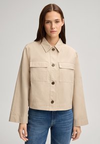 Giacca cropped beige con colletto, bottoni frontali e due tasche sul petto; realizzata in tessuto morbido con vestibilità rilassata.