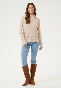 Pull en maille beige avec col roulé et motif à mailles ouvertes, associé à un jean bleu clair et des bottes marron à genoux.