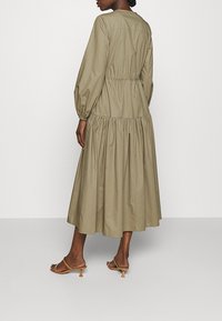Donna che indossa un lungo abito verde oliva con maniche lunghe e sandali con tacco a kitten beige, in piedi su uno sfondo neutro.