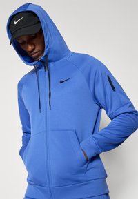 Mėlyna zip-up gobtuvinė su glotnia medžiaga, juoda Nike logotipas ant krūtinės, juodos raišteliai, šoniniai kišeniai ir užtrauktuku užsiseganti kišenė ant rankovės.
