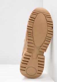Semelle d'une botte de randonnée beige avec une semelle profonde et large pour une bonne adhérence, reposant contre une surface blanche dans un environnement de studio.