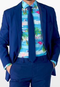 Traje azul marino con una corbata a juego, combinado con una camisa que presenta un estampado tropical de flamencos rosas y palmeras sobre un fondo azul claro.