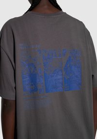 Szara bawełniana koszulka z luźnym fasonem, z niebieskim graficznym nadrukiem trzech map oraz szczegółowym tekstem. Okrągły dekolt, krótkie rękawy.