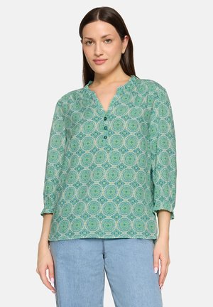 Femme portant un chemisier vert à manches longues avec un col à volants et un jean bleu, debout devant un fond blanc.