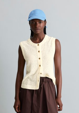 Femme portant un gilet tricoté crème sans manches boutonné, un pantalon marron foncé et une casquette de baseball bleu clair sur un fond gris uni.