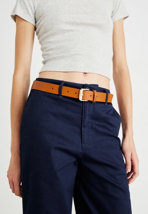 Καφέ δερμάτινη ζώνη με χρυσό μεταλλικό πόρπη, φορεμένη με σκούρα ναυτικά παντελόνια, δημιουργώντας έναν αντίθετο συνδυασμό χρωμάτων. Υφή στο φινίρισμα της ζώνης.