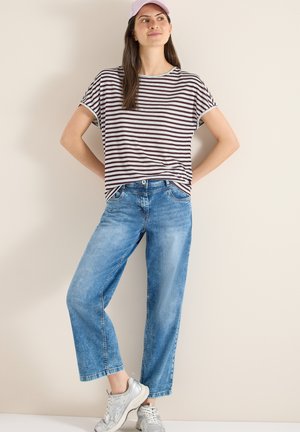 Frau mit rosa Mütze, gestreiftem Kurzarmshirt, blauen Jeans und weißen Turnschuhen, die mit den Händen in der Hüfte vor einer beigen Wand steht.