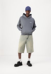 Grijze hoodie met voorzak, gedragen met losse lichte denim shorts. Zwarte chunky schoenen en een blauwe geribde beanie maken de outfit compleet.