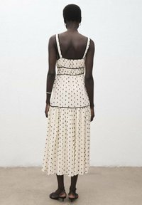 Vestido sin mangas de color crema con lunares negros, corsé ajustado, dobladillo evasé y ribetes negros en contraste. Diseño de espalda abierta, tejido fluido.