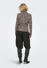 Femme aux cheveux blonds courts, portant une veste imprimé léopard, un pantalon noir ample et des bottes noires à talons hauts jusqu'aux genoux, debout de dos.