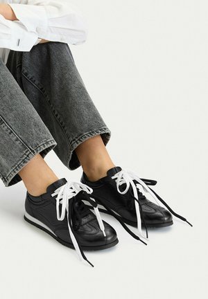 Person trägt schwarze Sneakers aus Leder mit ungleichfarbigen schwarzen und weißen Schnürsenkeln, kombiniert mit gekürzten dunkelgrauen Jeans und einem weißen Hemd.